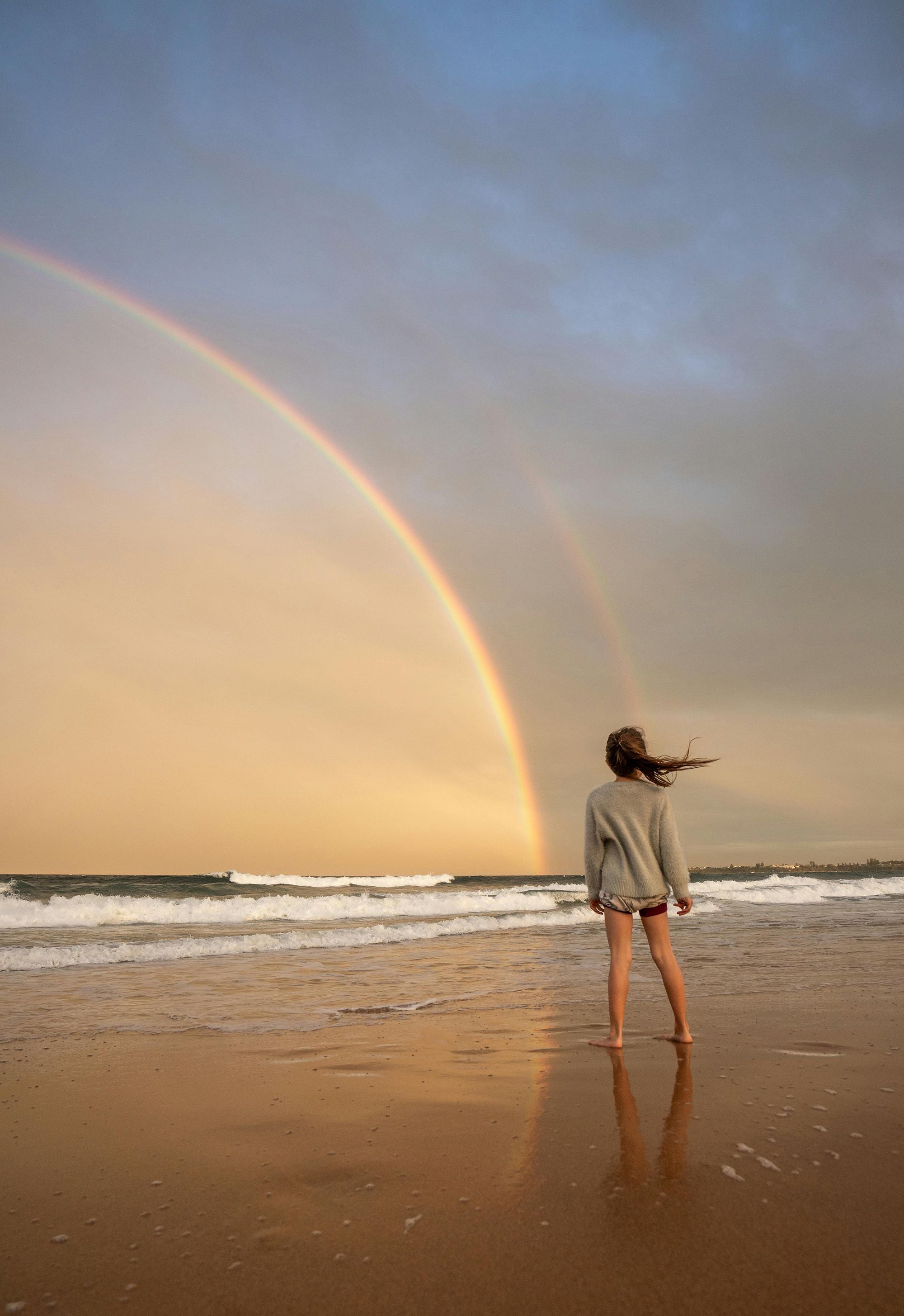 Image of a girl standing on the beach looking out over the sea at rainbow.