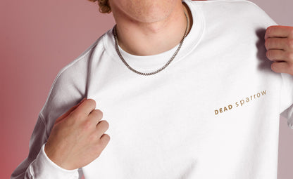 Close-up of a man wearing a white organic sweatshirt. Text "Deadsparrow" embroidered on the left side of the sweatshirt in gold thread. Product name Enough.