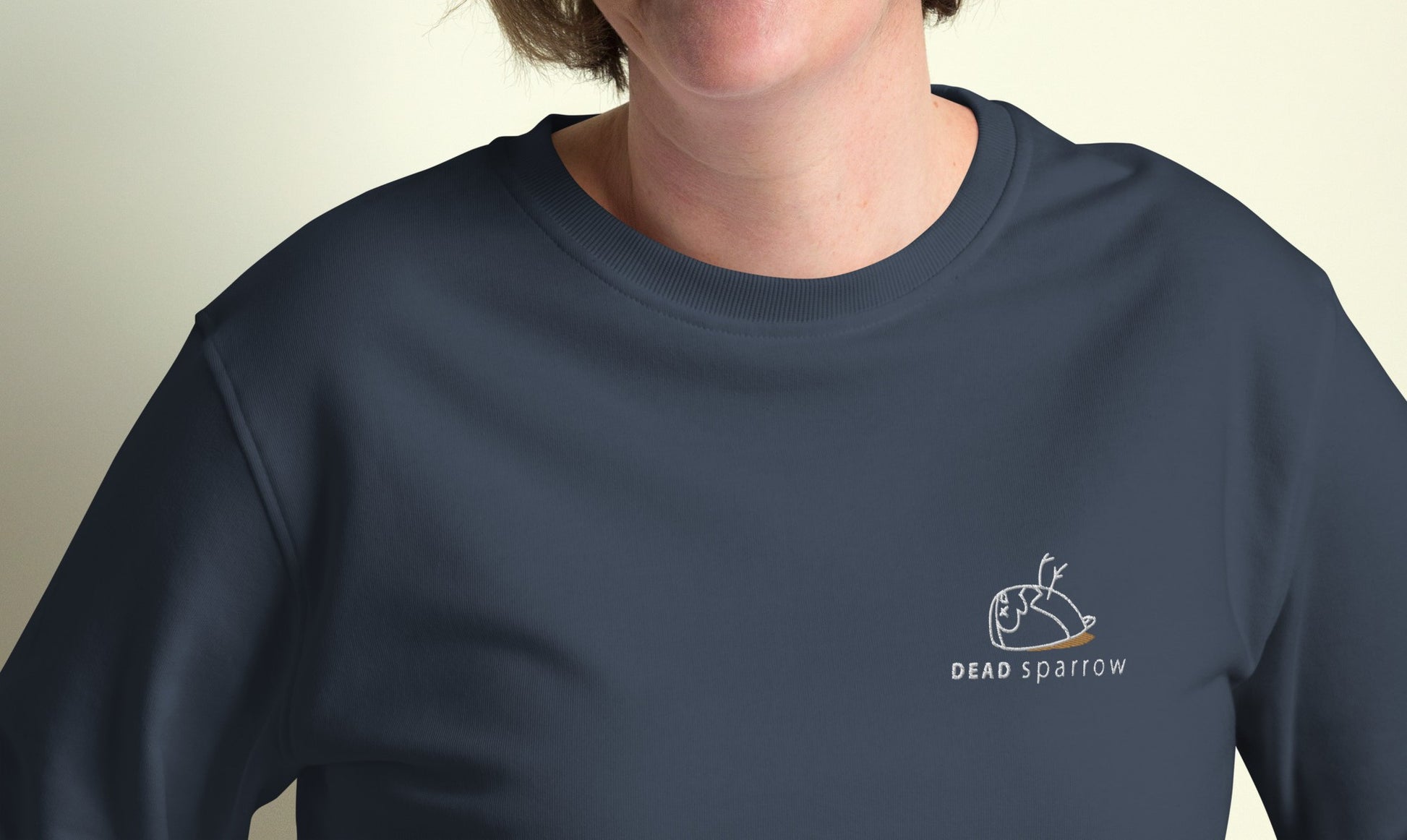 Close-up of a woman wearing an organic sweatshirt in the colour French navy. DEADsparrow logo embroidered on the left side in white and gold. Product name Significant. Significant is made from organic cotton and recycled materials