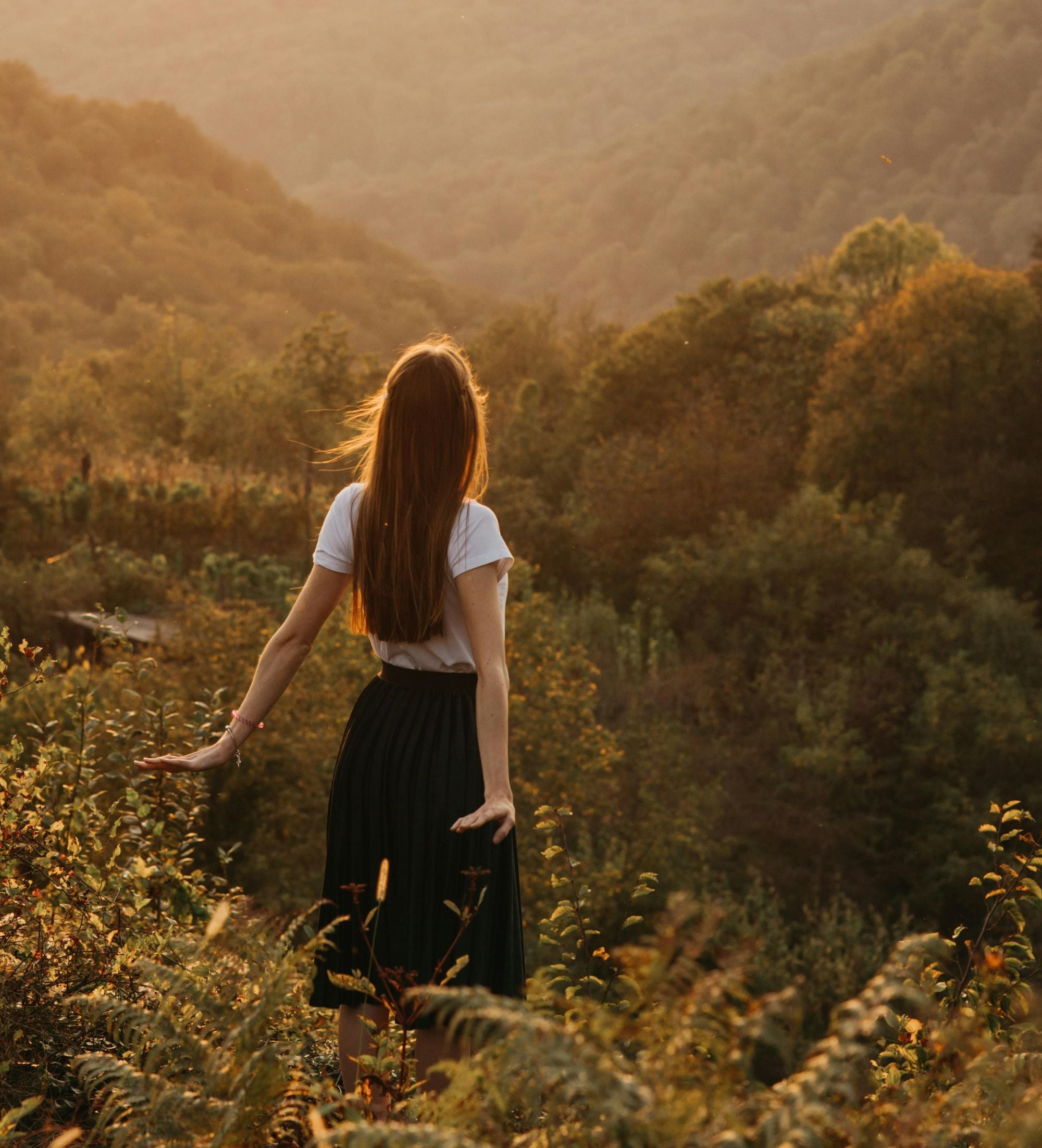 Woman wearing a white t-shirt and black skirt, looking out over the landscape during sunset.