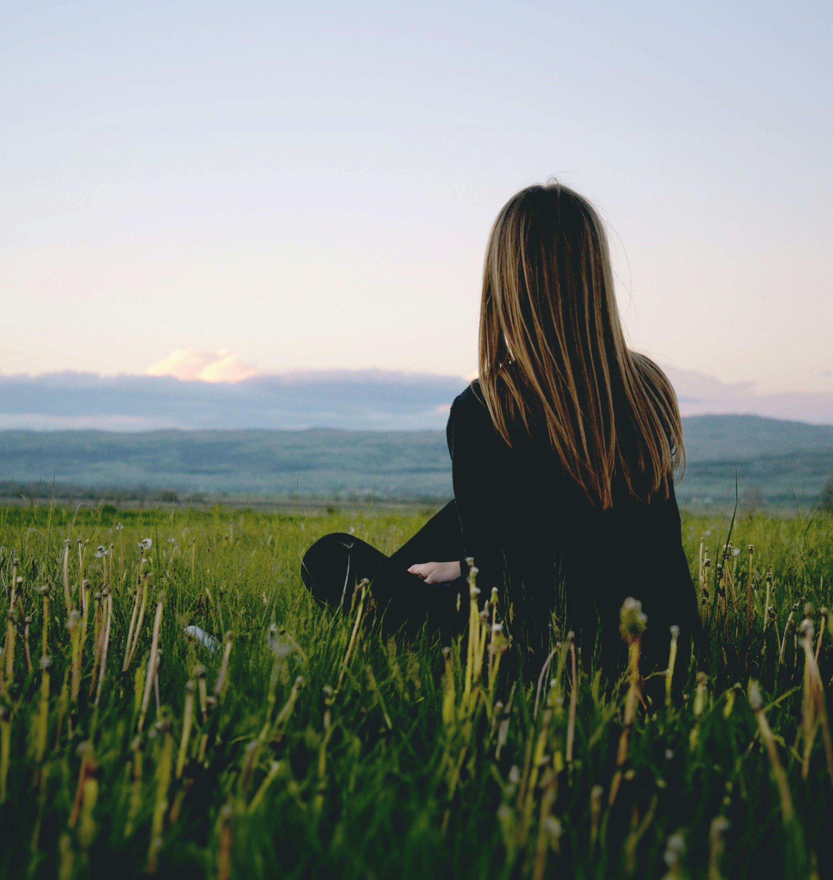 Woman sitting on the grass looking out over a landscape. Wearing black trousers and shirt.