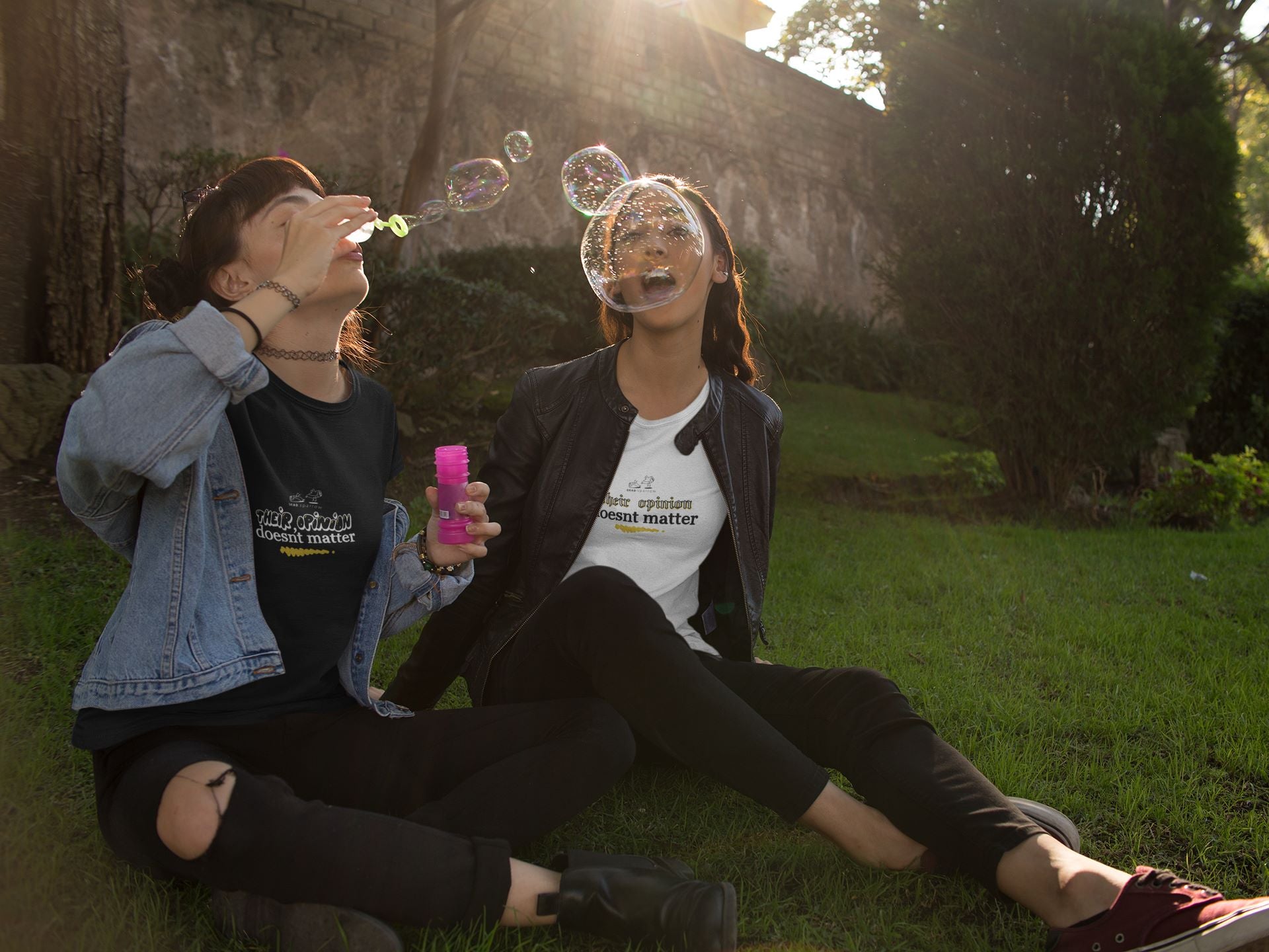 Picture of two women sitting outside on a sunny day, blowing bubbles and laughing. Both are wearing the "Nice" T-shirt, but in different colour prints.