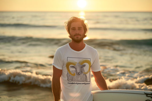 Image of a man holding a surfboard, with the sea behind him and it is sunset, wearing a white t-shirt with mouflon print. The product name is "Graceful". Below the print is the phrase "A symbol of grace and power".